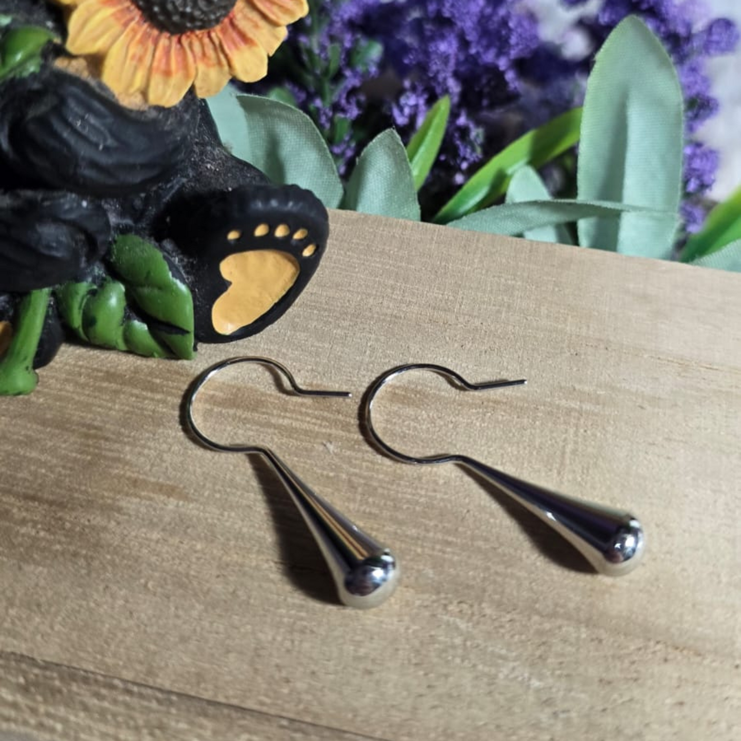 Par de aretes colgantes de acero inoxidable en forma de lágrima, diseño minimalista, sobre fondo rústico con flores decorativas.