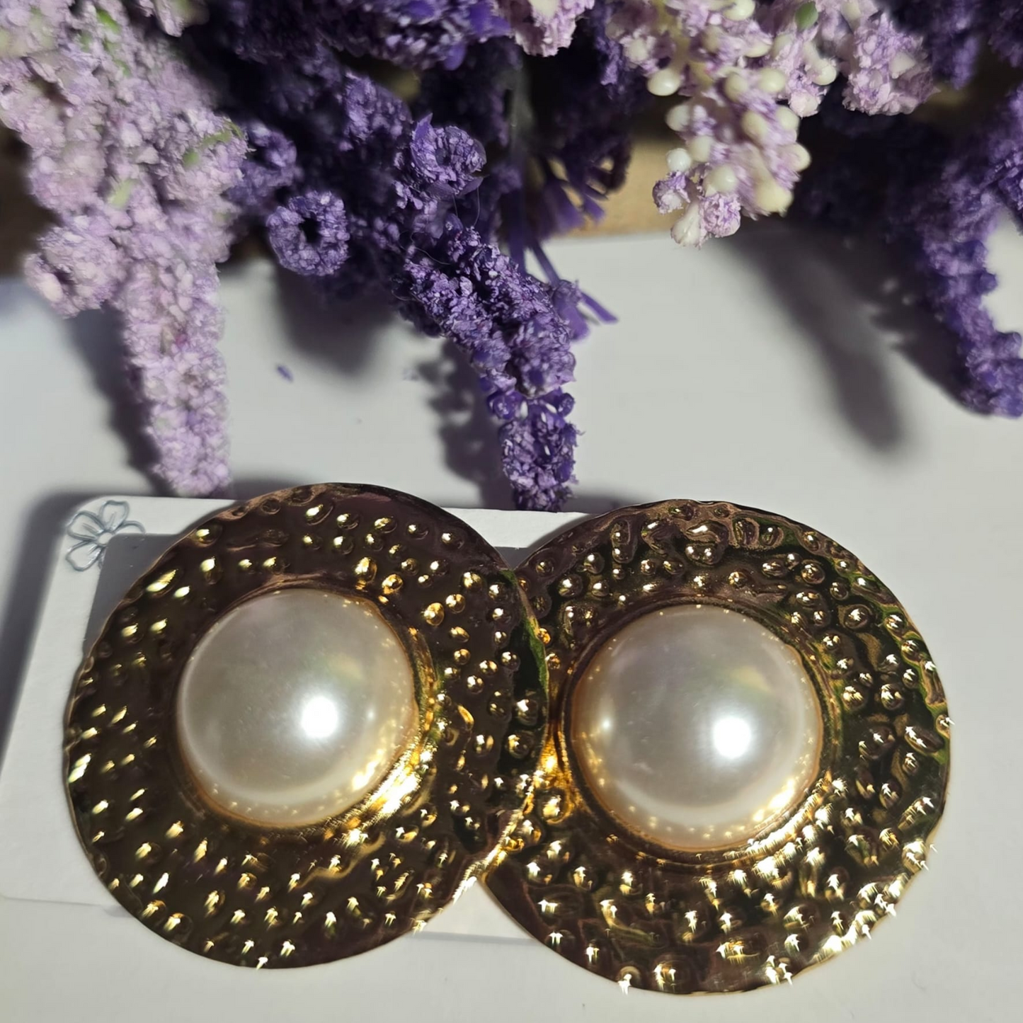 Aretes redondos dorados con textura y perla blanca central sobre fondo claro.