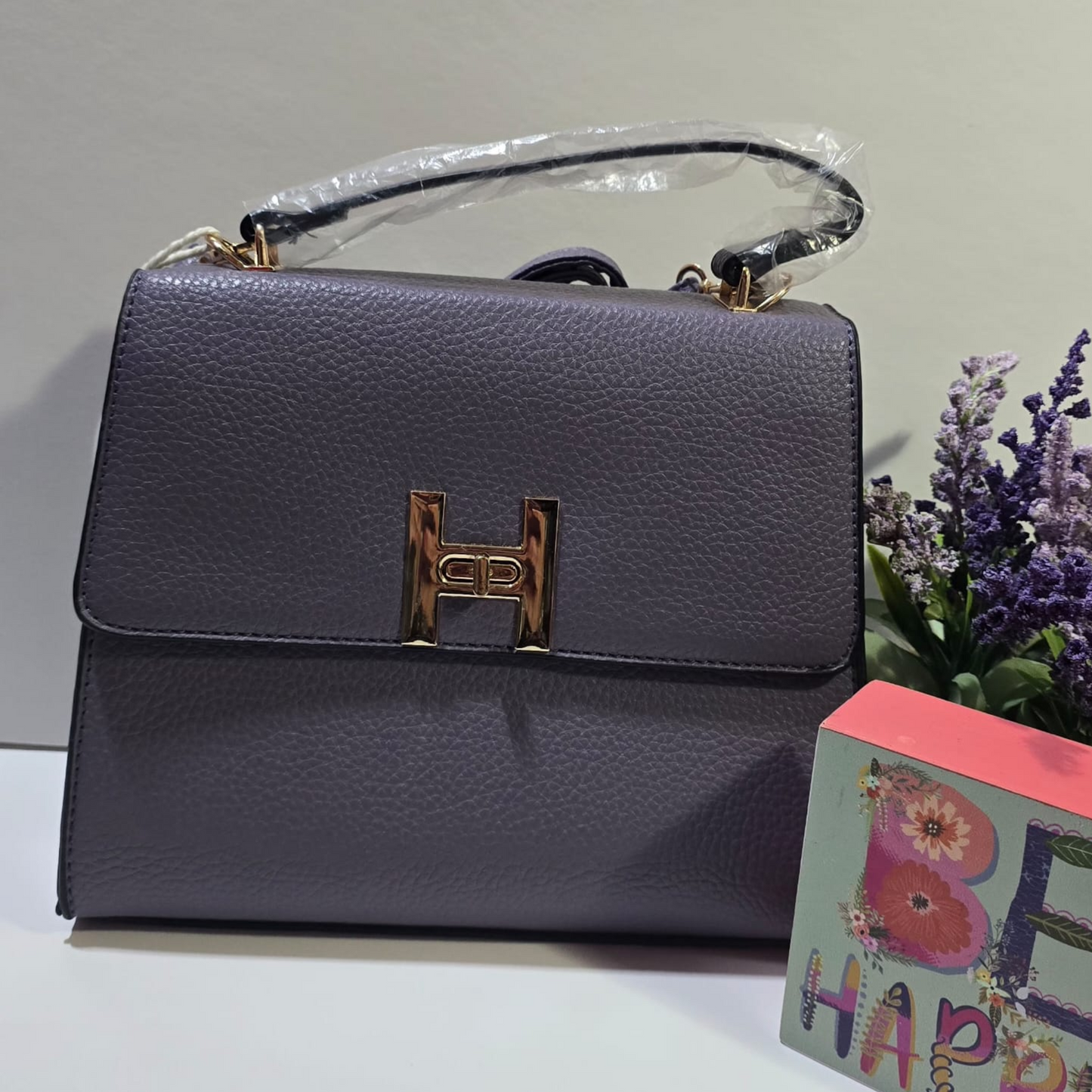 Cartera de cuero sintético color gris oscuro con hebilla dorada en forma de H y asa superior.