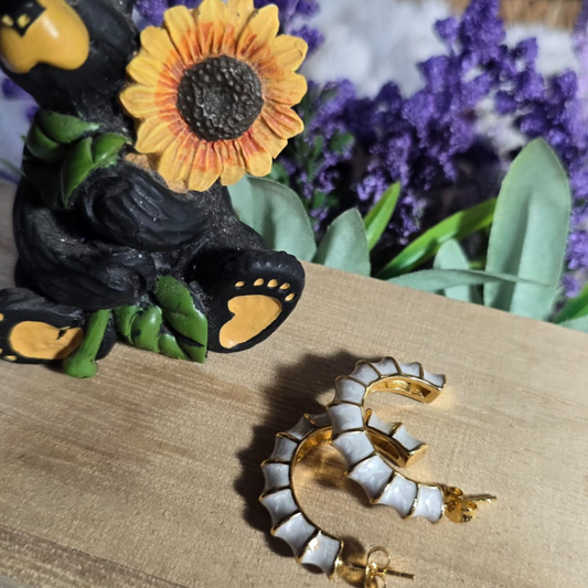 Aretes tipo aro dorado con relieves blancos colocados sobre una base de madera, con fondo decorativo de oso negro con girasol y flores moradas.