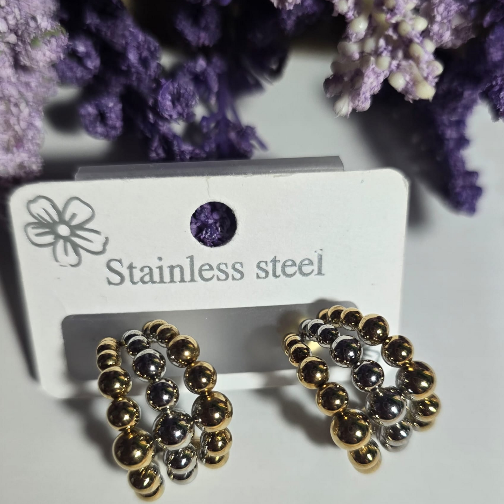 Aretes de acero inoxidable bicolor con diseño de esferas doradas y plateadas entrelazadas, exhibidos sobre fondo blanco con flores lavanda.

