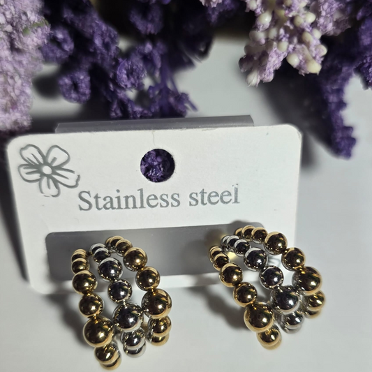 Aretes de acero inoxidable bicolor con diseño de esferas doradas y plateadas entrelazadas, exhibidos sobre fondo blanco con flores lavanda.

