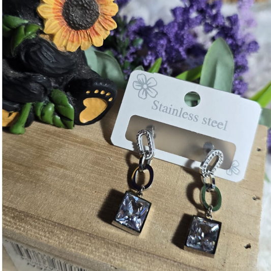 Aretes colgantes de acero inoxidable con eslabones decorativos y dije cuadrado brillante, presentados sobre una base de madera junto a una figura de oso con girasol y flores moradas.