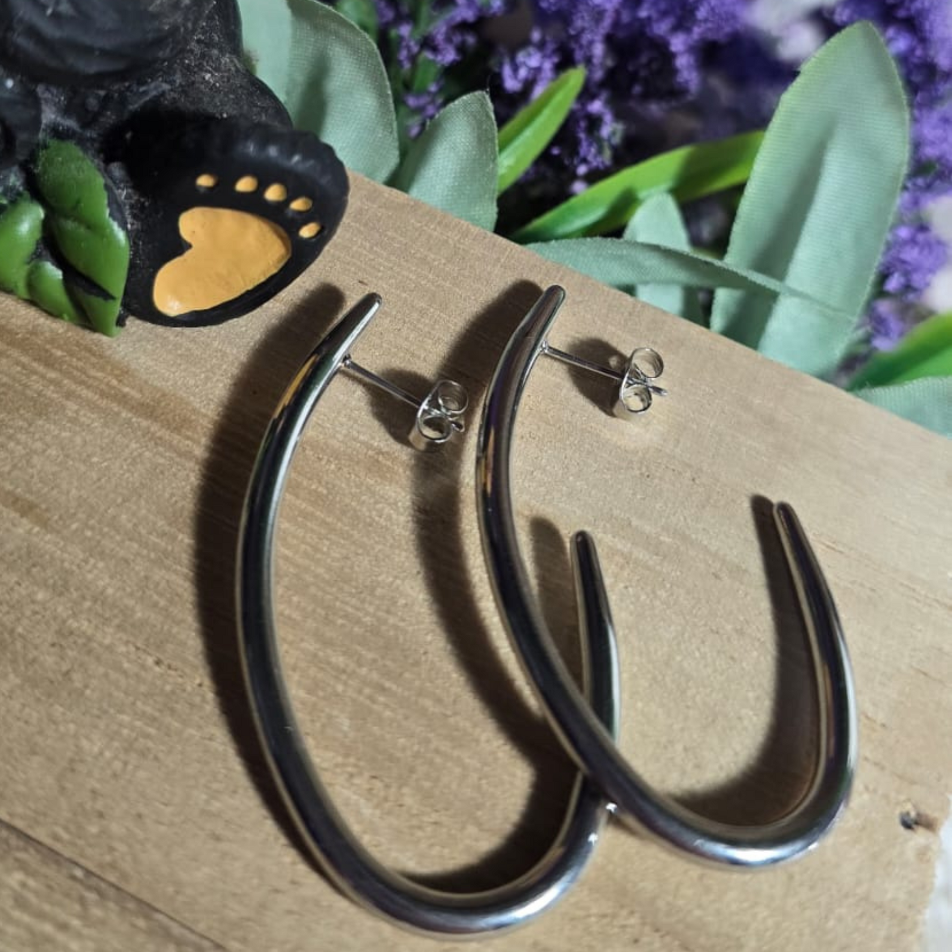 Aretes curvos y alargados de acero inoxidable sobre una base de madera, acompañados por una figura de oso con girasol y fondo floral violeta.