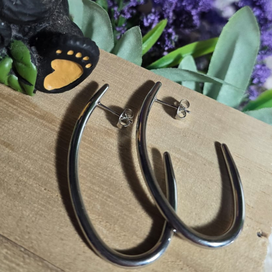 Aretes curvos y alargados de acero inoxidable sobre una base de madera, acompañados por una figura de oso con girasol y fondo floral violeta.