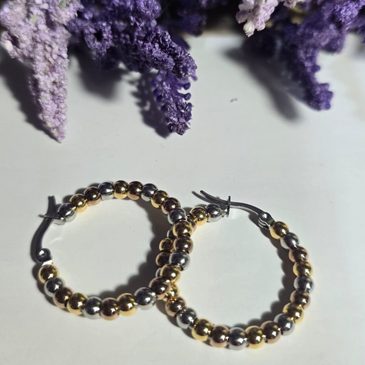 Aretes de aro con bolitas metálicas en tonos dorado y plateado sobre fondo claro.

