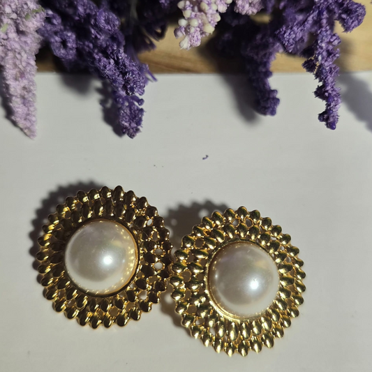 Aretes dorados circulares con diseño texturizado y perla blanca central sobre fondo claro.

