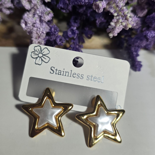 Aretes en forma de estrella con borde dorado y centro nacarado, fabricados en acero inoxidable, colocados sobre fondo blanco con flores lavanda.

