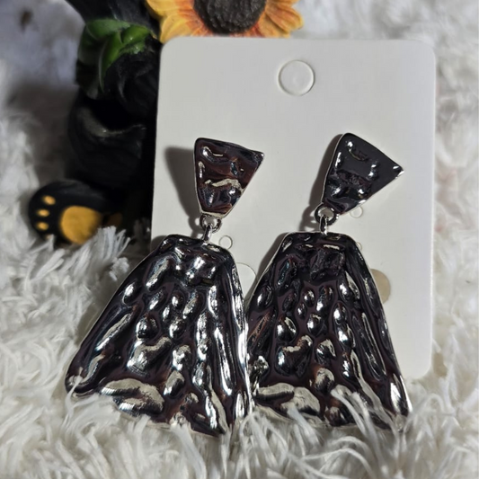 Aretes plateados grandes con diseño geométrico triangular y textura martillada, presentados sobre fondo blanco peluche junto a decoración de girasol y figura de oso.
