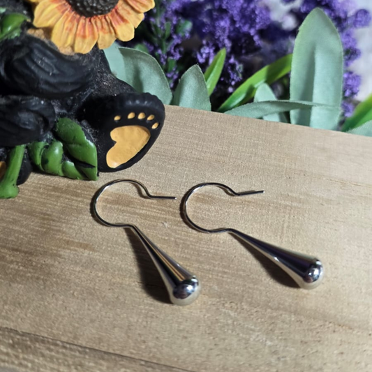 Par de aretes colgantes de acero inoxidable en forma de lágrima, diseño minimalista, sobre fondo rústico con flores decorativas.
