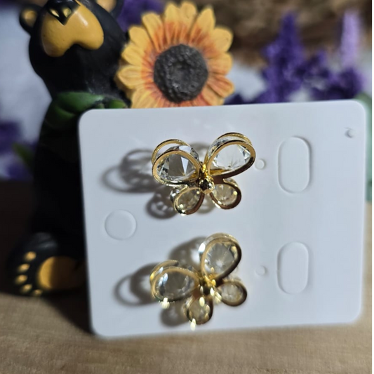 Aretes dorados en forma de mariposa con cristales transparentes, presentados sobre fondo decorativo con girasol y figura de oso.
