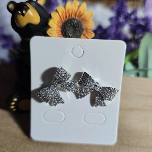 Aretes en forma de moño plateado con incrustaciones brillantes, colocados sobre tarjeta blanca y decorados al fondo con girasol, oso decorativo y flores moradas.