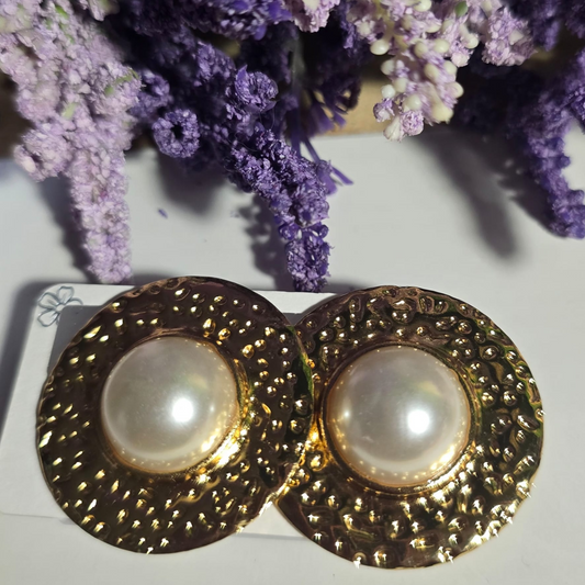 Aretes redondos dorados con textura y perla blanca central sobre fondo claro.

