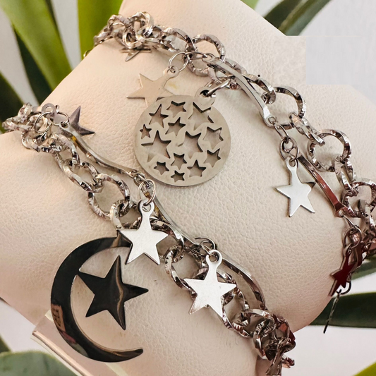 Pulsera plateada de acero inoxidable con dijes colgantes de luna y estrellas sobre un cojín blanco de exhibición.