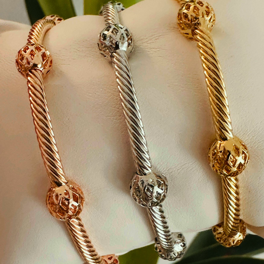 Tres pulseras rígidas tipo cable en dorado, plateado y oro rosa, con esferas texturizadas, exhibidas sobre un soporte blanco.