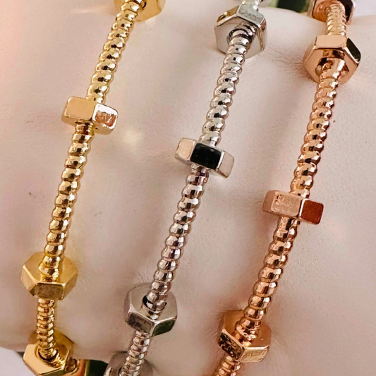 Tres pulseras rígidas tipo tuerca con textura estriada en dorado, plateado y oro rosa, exhibidas sobre un soporte blanco.