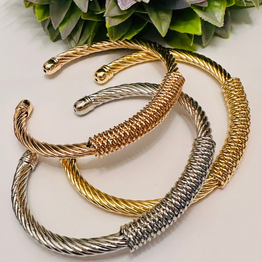 Tres pulseras rígidas abiertas de acero inoxidable en colores oro, plata y oro rosa, con diseño de cable torcido y centro envuelto en alambre, sobre fondo blanco
